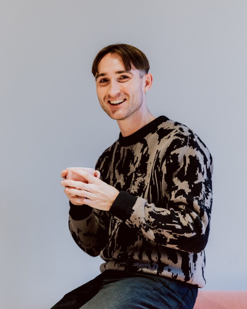 Ben sitting on a pink sofa with a cup f coffee smiling into the camera.