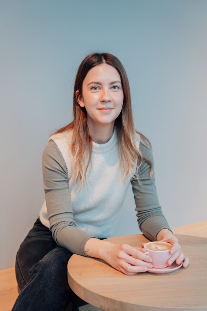 Melissa sitting at a table with a cup of coffee smiling into the camera.